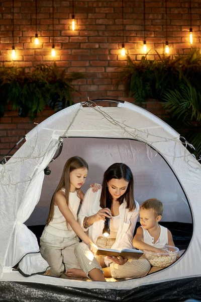 Mom reads children a bedtime story sitting in a tent at home. Mother son and daughter hug and read a book with a flashlight in their hands
