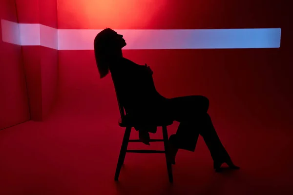 Woman on red background sits on chair, beam of light behind her face. Sexy and confident woman in black jacket