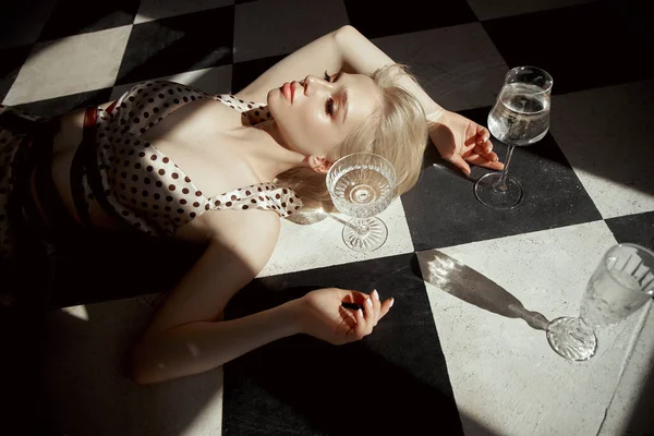 手里拿着玻璃杯的女人躺在地板上 一个老旧的内饰 浪漫的金发姑娘 女性身体上的窗户阴影 — 图库照片