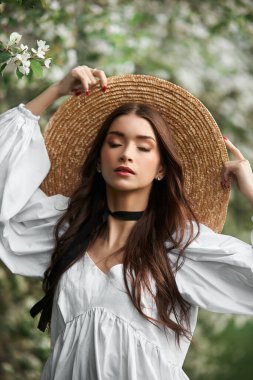 Kahverengi saçlı, büyük hasır şapkalı ve beyaz elbiseli bir kadın çiçek açan beyaz ağaçların arkasında poz veriyor. Romantik görünüş, doğal güzellik, temiz yüz derisi.
