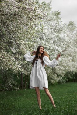 Hasır şapkalı beyaz elbiseli genç bir kadın baharda çiçek açan bahçe parkında yürüyor. Bahar geldi, romantik ruh hali.
