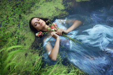 Güzel, romantik bir kadın, bataklıkta, mavi, uzun elbiseli, çiçekli bir şekilde yatıyor. Yüzeysel esmer, şeffaf elbiseli, bataklık çamur ördek otlu. Kitap kapağı