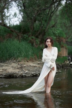 Uzun beyaz elbiseli erotik genç bir kadın nehir kenarındaki bir bahar parkında yürüyor. Seksi esmer doğada poz veriyor.