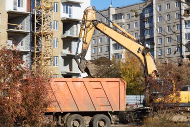 excavator is seen removing dirt from a construction area and loading it into a large orange truck. setting features surrounding buildings and autumn foliage under clear skies.