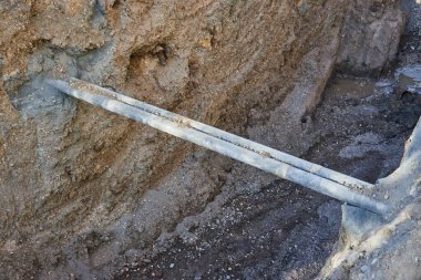Workers are repairing utility pipes located in a deep trench at a construction site. ground is uneven, with dirt and gravel around the pipes under clear blue skies.