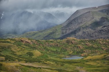 beautiful valley displays vibrant green vegetation and a calm lake, nestled among majestic mountains. Clouds drift across the sky, enhancing the natural scenery. Russia, Altai Mountains