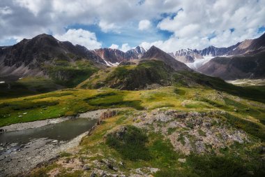 Lush green valley flanked by rugged mountains displays a serene river winding through the landscape under a partly cloudy sky. Nature thrives in this untouched wilderness. Russia, Altai Mountains