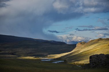 İçinde dolambaçlı bir nehir, kayalık kayalıklar ve uzak dağların bulunduğu huzurlu bir vadinin üzerinden altın ışık süzülür. Kara bulutlar öğleden sonra manzaraya karşı çarpıcı bir kontrastlık yaratır..