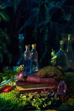Colorful potions sit on an old book, surrounded by green moss and stones in a dark forest. Smoke rises, adding a magical touch to the scene.