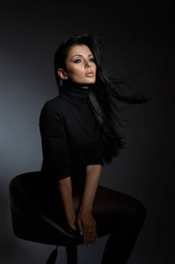 confident woman sits gracefully on a stool, her long hair flowing softly. dramatic lighting emphasizes her features, creating a captivating atmosphere in the studio.