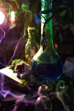 glowing blue potion in an ornate bottle sits among mystical items. Soft smoke rises, and other vintage bottles create an intriguing and mysterious atmosphere in a dark setting.