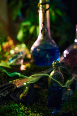 Bottles of blue potions rest on mossy ground in a forest. Soft light creates a magical atmosphere as nearby artifacts glimmer. scene is enchanting and evocative of fantasy tales.