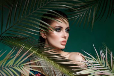 model showcases striking makeup and fashion while posing amongst lush green palm leaves. vibrant backdrop highlights her features in a creative studio setting.