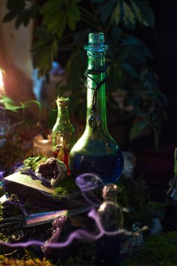 Renkli iksir şişeleri, kristallerle çevrili yosunlu bir masanın üzerinde ve hafifçe aydınlatılmış bir odada mistik eşyalar bulunmakta. Titrek ışık sihirli bir atmosfer ekler..