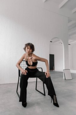 Siyah kıyafetli bir model minimalist bir stüdyoda cesur bir poz sergiliyor. Beyaz duvarlar ve çağdaş tasarım öğeleri yer almaktadır, moda açıklamasını geliştiren.