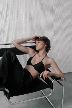 Siyah bikinili bir kadın ve pantolonu tarafsız bir arka planı olan parlak bir sandalyede dinleniyor. Güneş ışığı içeri süzülüyor, sakin ifadesini ve şık duruşunu vurguluyor.