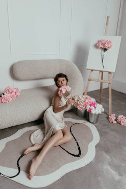 Model zarif bir şekilde stüdyoda renkli çiçekler taşıyan eşsiz bir koltuğa oturur. Yakınlardaki sehpada çiçek sergileniyor. Yumuşak ışıklandırılmış çevrenin sanatsal havasını geliştiriyor..