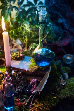 Parlak mavi iksirle dolu cam şişe ahşap bir yüzeye oturur. Etrafı yosun, mum ve renkli dumanla çevrilidir. Atmosfer büyüleyici ve büyüleyici..