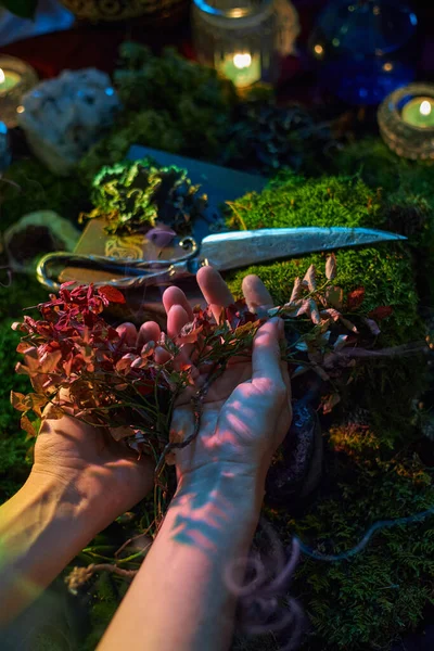 Ellerinde mumlar ve yosunlarla çevrili kurumuş kırmızı otlar var. Gümüş makas yakınlarda duruyor, alacakaranlıkta büyülü bir ortam yaratıyor..