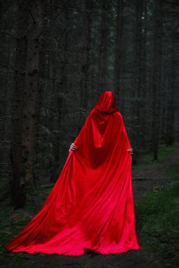 Dalgalı kırmızı pelerin giyen bir figür sık bir ormanda dar bir yolda duruyor. Sahnenin etrafını saran uzun ağaçlar ve ışık loş, gizemli bir atmosfer yaratıyor..