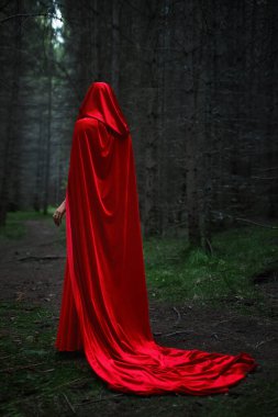 Dalgalı kırmızı bir pelerinle sarılı bir figür sık, gölgeli bir ormanda dar bir patika boyunca gezinir. Sabah ışığı kalın ağaçların arasından geçerek ürkütücü bir atmosfer yaratıyor..