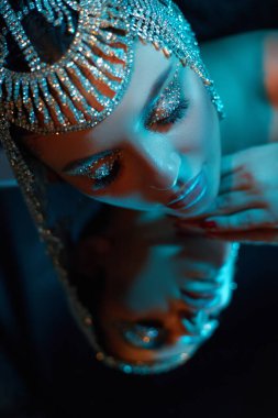 Detaylı başlıklı model, parlak makyajla kafasında dinleniyor. Yumuşak ışık, onun güzelliğini ve yansımasını vurgulayan sakin bir atmosfer yaratır..