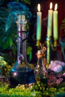Mavi sıvıyla dolu iki cam şişe, mum ve bitkilerle çevrili yosunlu zeminde oturur. Duman havada süzülür, büyüleyici bir atmosfer yaratır..