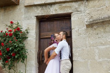 Kırmızı güllerle çevrili taştan bir duvarın yanında mutlu bir şekilde kucaklaşırlar. Özel bir an için giyinmişler, aşk ve mutluluk yayıyorlar..