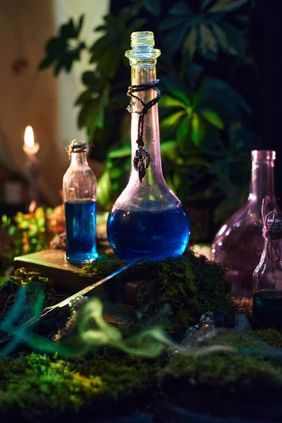 Parlak mavi sıvı içeren çeşitli cam şişeler yemyeşil yosunlarla kaplı bir yüzeye yerleştirilmiştir. Atmosfer mum ışığıyla hafifçe aydınlatılır, doğal çevreyi ön plana çıkarır..