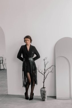 Model, püsküllü, şık bir siyah elbisenin içinde güvenle duruyor. Minimalist stüdyonun şık tasarım ve dekorasyon elemanlarını vurgulayan yumuşak renkleri var.