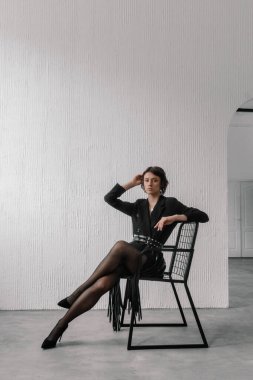 Siyah elbiseli bir kadın zarif bir şekilde parlak, minimalist bir odada oturuyor. Duruşu özgüven ve zarafet yayıyor, modern moda ve tasarım sergiliyor..