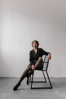 Genç bir kadın modern bir stüdyoda zarif, siyah bir sandalyede poz veriyor. Giysileri şık ve ışıklandırma minimalist ortamı güçlendiriyor, çarpıcı bir görünüm yaratıyor..