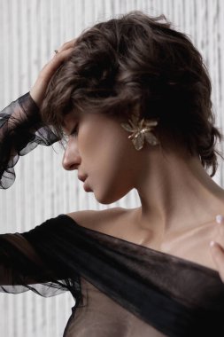 Yumuşak ışıklı bir ortamda, kısa saçlı bir kadın zarif bir poz verir, siyah bir bluz giyer ve zarif ve çekici bir tavırla karışık küpelerini gösterir..