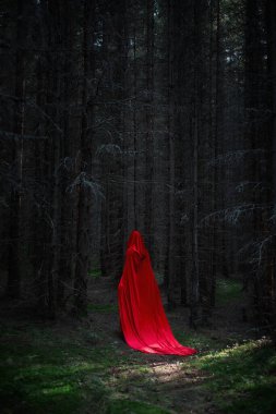 Akan kırmızı bir pelerinle örtülü figür, alacakaranlıkta uzun ve karanlık ağaçların arasında sessizce hareket eder. Gölgeler görünür, esrarı arttıran ürkütücü bir atmosfer yaratır..