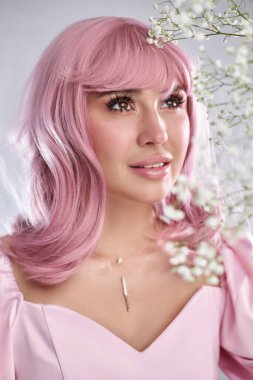 Pembe saçlı bir kadın, narin beyaz çiçeklerle çevrili iken nazikçe poz verir. Güzelliğini arttıran hafif ışıklı bir ortamda sakin bir yüz ifadesi sergiler..