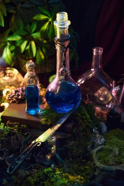 Parlak mavi sıvılar ahşap bir yüzeyde çeşitli cam şişelerde görülür. Şişelerin etrafı yosun ve bitkiler gibi doğal elementlerle çevrili..