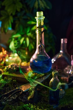 Mavi sıvıyla dolu birkaç cam iksir şişesi ahşap bir masada oturur. Masa, düşük ışık altında bitki ve otlarla kaplı. Gizemli bir sahne yaratıyor..