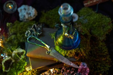 Mavi cam şişe, metalik bir bıçak ve taşın yanında yosunlu bir yüzeye oturur. Arka planda küçük ışıklar parlıyor, geceleri mistik bir atmosfer yaratıyor..