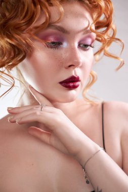 Kıvırcık kızıl saçlı manken stüdyoda poz verirken renkli göz makyajı ve koyu renk ruj yapıyor. Işıklar yüzüne hafifçe dokunduğunda yüzünün rengini ortaya çıkarıyor..