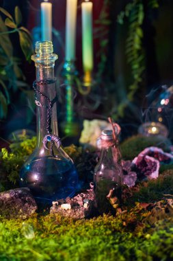 Sıvı dolu iki küçük cam şişe yeşil yosun üzerine yerleştirilir. Yakındaki mumlar yanıyor ve bölgeden duman yükseliyor. Gizemli bir atmosfer yaratıyor..