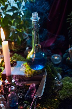 Mavi sıvıyla dolu cam şişe bir mum, bitkiler ve dekoratif eşyalarla çevrili ahşap bir masanın üzerinde duruyor. Sıcak akşam ışığında duman yükselir mistik bir sahne yaratır..