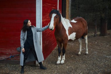 Kadın kırmızı bir ahırın yanında kahverengi ve beyaz bir atın yanında duruyor. Kırsal kesimde gülümserken ata nazikçe dokunur. Ağaçlar arka planda yumuşak bir ışığın altında görünür..