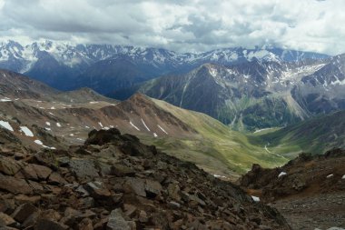 Tepeler ve vadiler manzara boyunca uzanırken bulutlar gökyüzünü kaplıyor. Dağlar gün ışığında arka planda yükselirken, Rocky arazisi ön planda görülebilir..