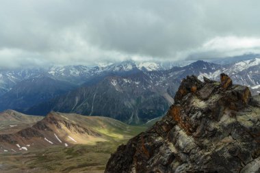 Bulutlar gökyüzünü kaplarken dağ sıraları uzak mesafelere kadar uzanıyor. Kayalık ön plan yeşil tepelere ve gri gökyüzünün altındaki karlı tepelere karşı duruyor..