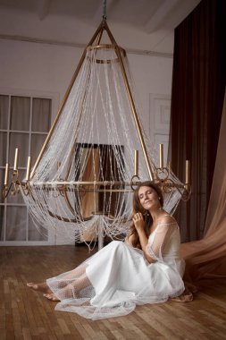 Kadın ahşap bir zeminde, üzerinde büyük bir avize olan bir odada oturuyor. Yumuşak ışıklandırma boşluğu doldurur dizine yaslandığında, rahatlamış ve düşüncelere dalmış görünür..