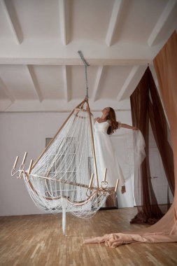 Beyaz elbiseli dansçı, büyük bir avizeye asılı dururken zarifçe hareket ediyor. Odanın ahşap zeminleri ve çeşitli renklerde kumaş perdeleri var..