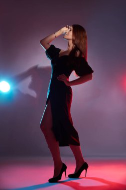 Kadın stüdyo ortamında duruyor, siyah bir elbise ve yüksek topuklularla poz veriyor. Renkli ışıklar yukarı baktığında canlı bir atmosfer yaratıyor..