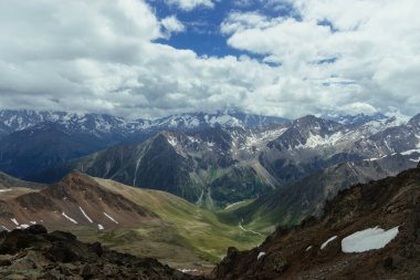 Dağların manzarası yeşil vadiler ve kayalık tepelerin karışımıyla manzara boyunca uzanıyor. Bulutlar gökyüzünde hareket ediyor, araziye gölge düşürüyor. Öğlen vakti yaz geldi..