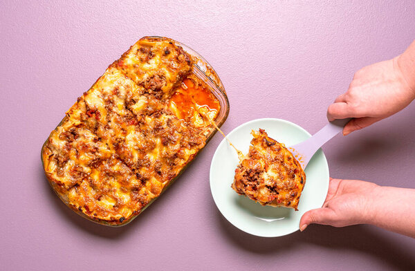 Above view with woman hands serving on a plate a portion of freshly-baked lasagna. Delicious homemade lasagna top view on a purple background.