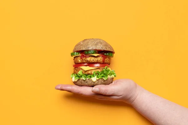Turuncu arka planda ev yapımı vejetaryen burger tutan bir kadın eli. Soya köfteli sebze burger, vejetaryen kaju peyniri, remoulade sosu ve sebze.